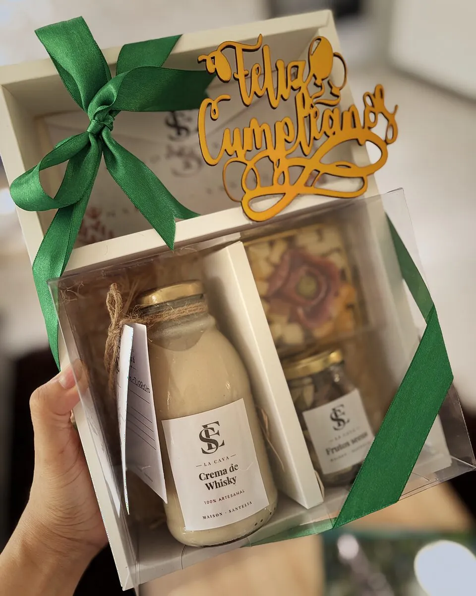 Caja de cumpleaños de La Cava sostenida en la mano, con crema de whisky, frutos secos, quesos y una cinta verde